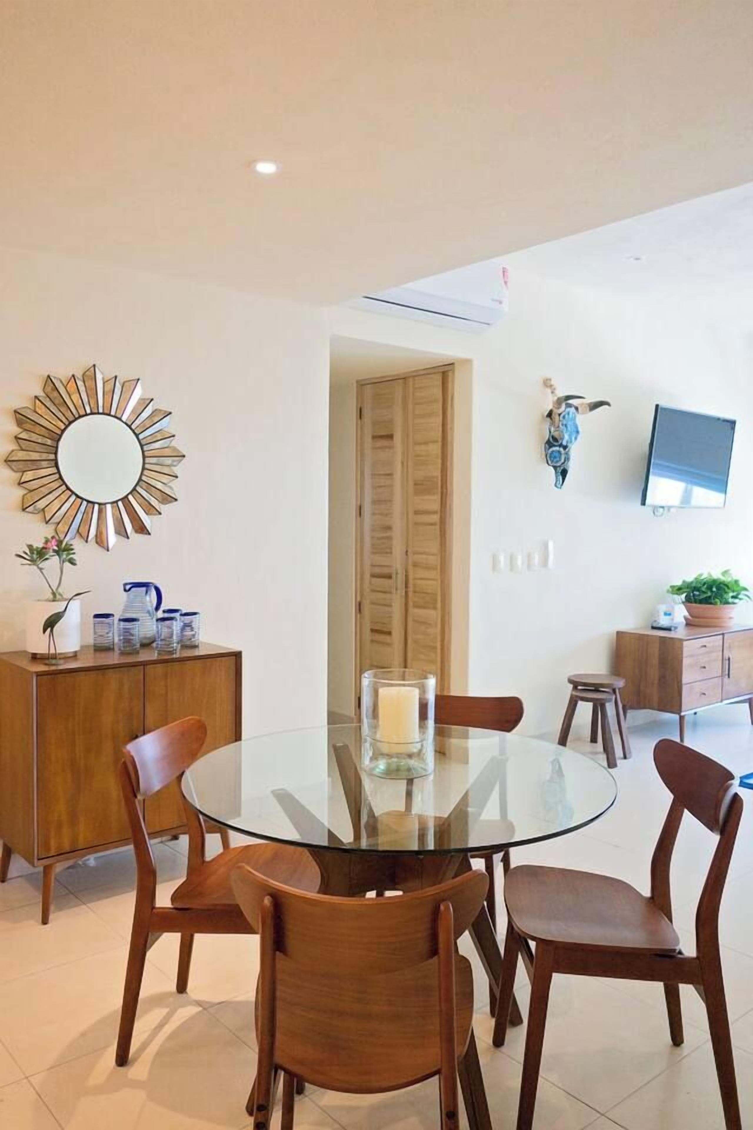 A dining area with a glass table, wooden chairs, a decorative mirror, and a television mounted on the wall.