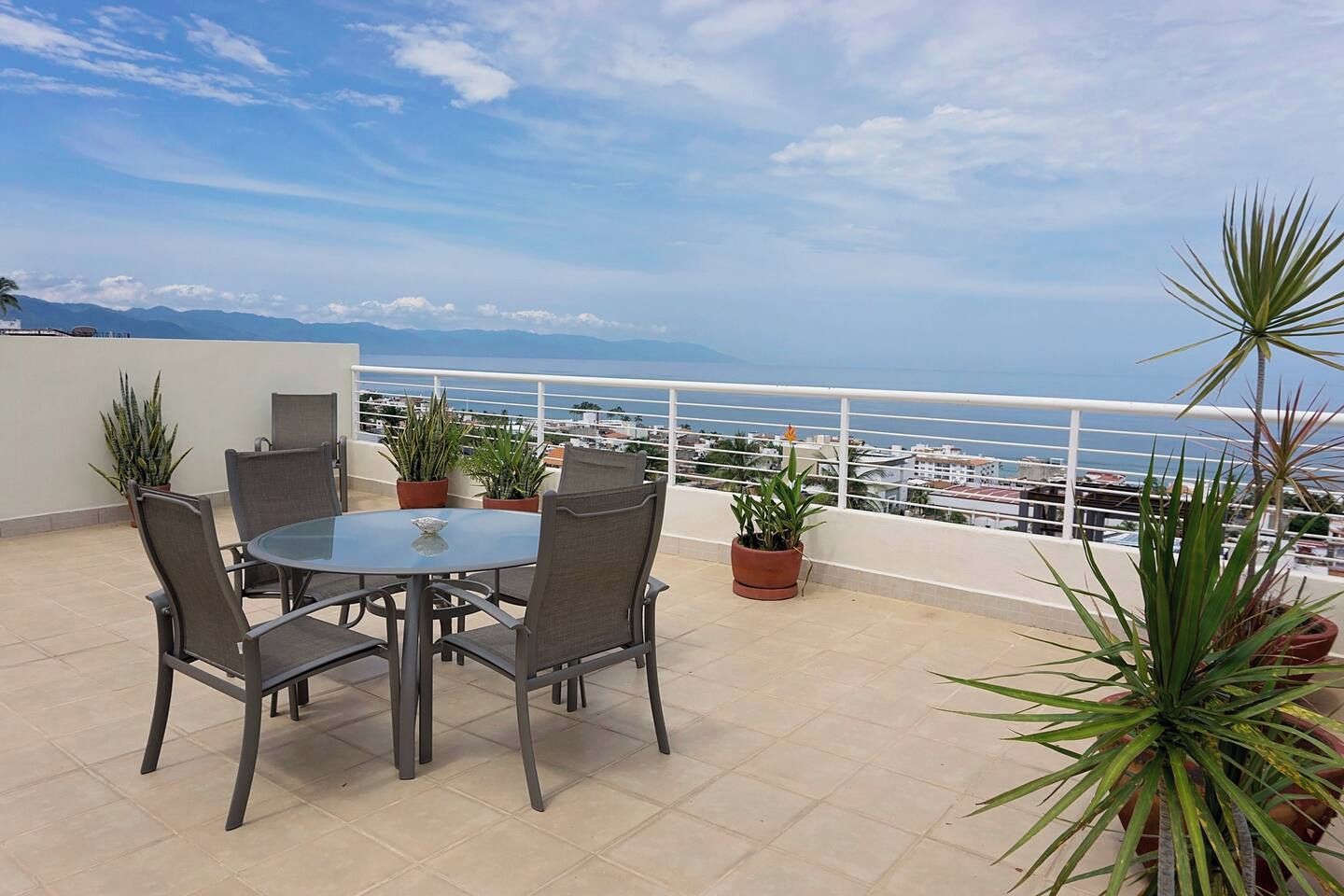 A terrace with a round table and chairs, surrounded by potted plants, overlooking a coastal view.