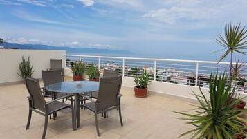 A terrace with a round table and chairs, surrounded by potted plants, overlooking a coastal view.