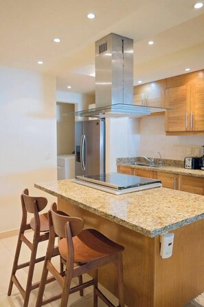 A modern kitchen with a stainless steel refrigerator, granite countertop, wooden cabinets, and a built-in island with bar stools. - Beautiful, Airy Condo Near Boardwalk**** (Puerto Vallarta)