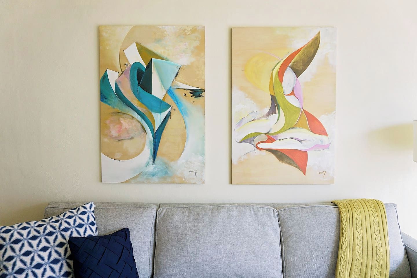 Two abstract paintings on a wall above a grey sofa with patterned cushions.