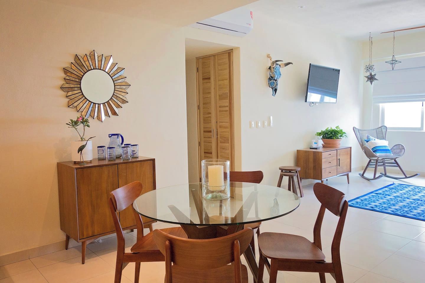 A modern dining area with a glass table, wooden chairs, a decorative mirror, a television, and a potted plant.