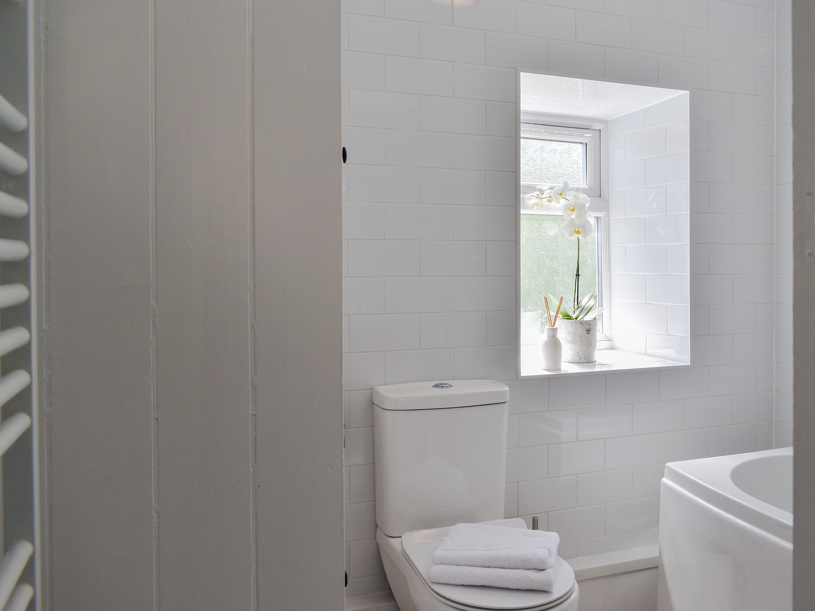 A bathroom with white tiles, a toilet, a sink, and a window with flowers.