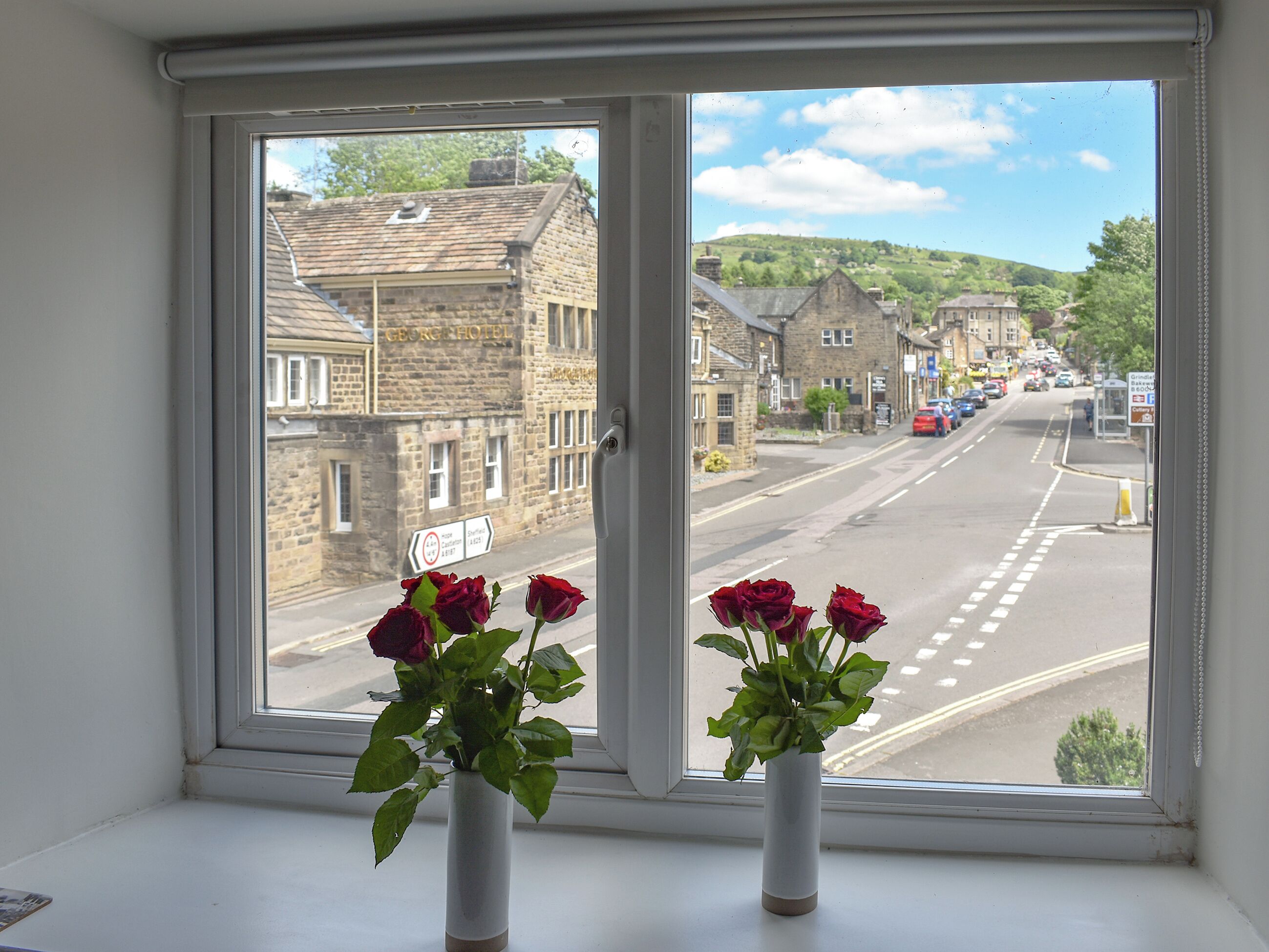 A window with a view of a street, a building labeled 'GEORGE HOTEL,' and two vases with flowers.