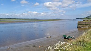 A riverbank with a green boat tied to a dock, surrounded by grassy areas and wildflowers. - 2 bedroom accommodation in Newburgh, near Cupar (Newburgh, near Cupar)