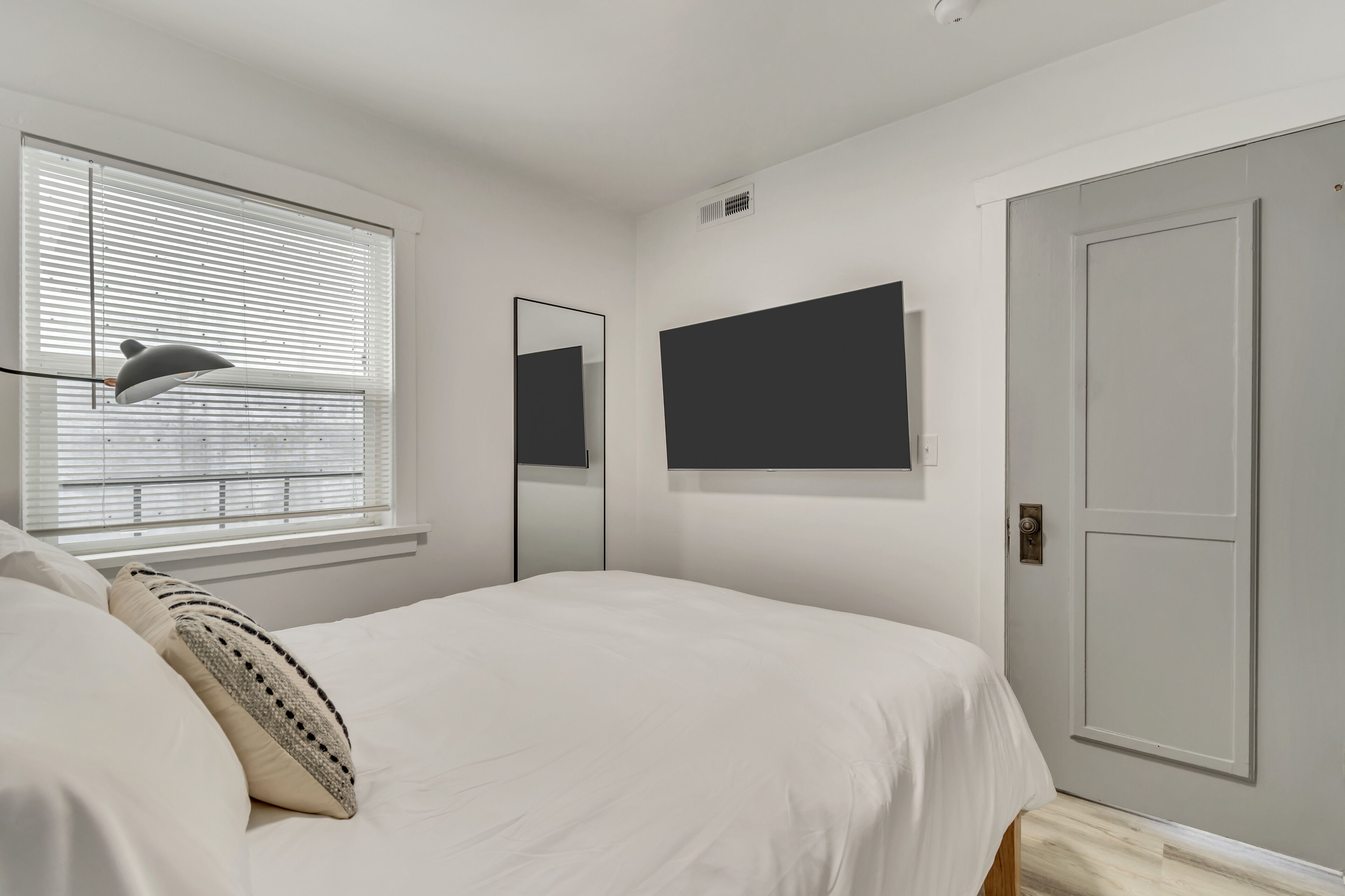 A bedroom with a bed, a bedside lamp, a window with blinds, a wall-mounted TV, and a door.