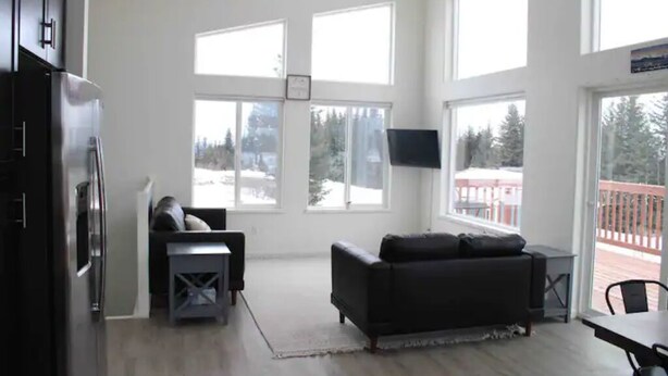 A spacious living area with a dark-colored sofa, a flat-screen TV mounted on the wall, and large windows offering a view of a snowy deck outside. - Peaceful Homer Getaway (Homer)