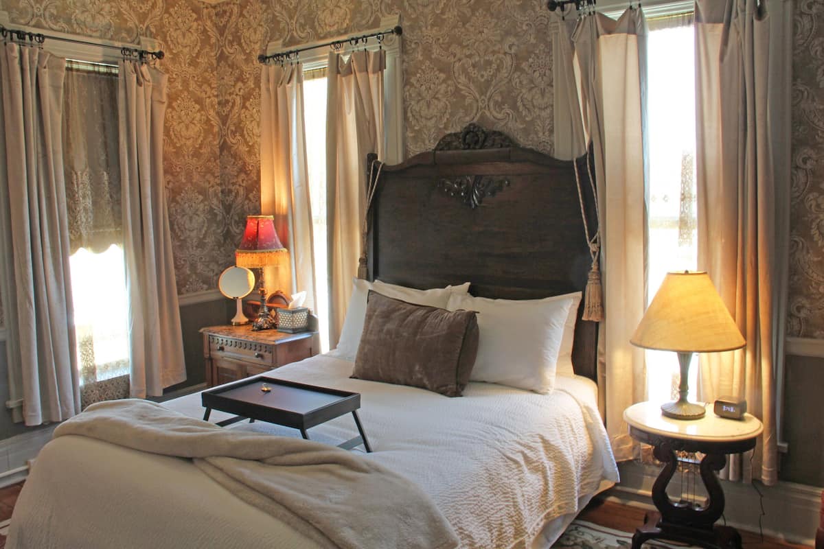 A bedroom with a large bed, a bedside table with a lamp, and curtains on the windows.