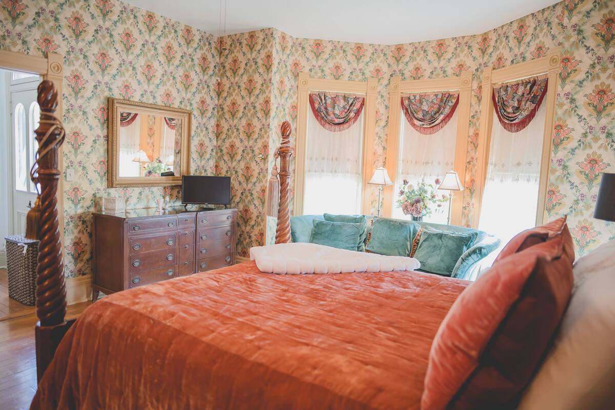 A bedroom with floral wallpaper, a wooden bed with a red bedspread, a dresser, a mirror, and two windows with curtains.