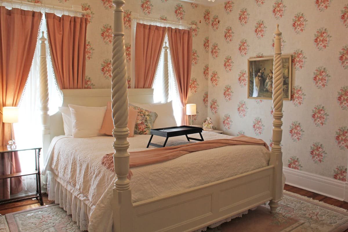 A four-poster bed with a canopy, a bedside table with a lamp, floral wallpaper, and a framed picture on the wall.