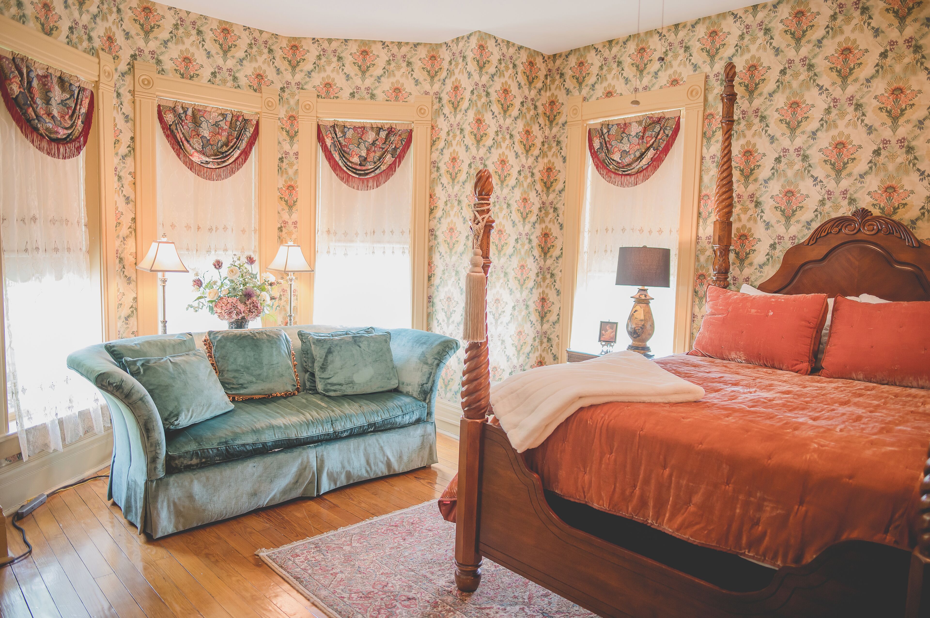 A room with a wooden bed, a green sofa, floral wallpaper, and a large window with curtains.