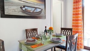 Une table à manger dressée avec des assiettes, des verres et une bouteille, un tableau représentant un bateau accroché au mur et un rideau orange.