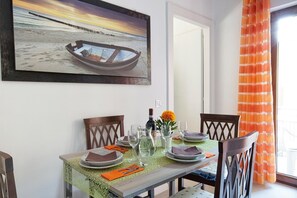 Une table à manger dressée avec des assiettes, des verres et une bouteille, un tableau représentant un bateau accroché au mur et un rideau orange.