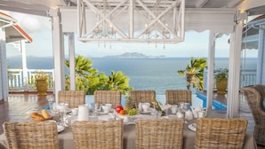 A dining area with wicker chairs and a table set for a meal, overlooking a body of water and mountains. - Luxury Villa in the French Carribean with a stunning view (Vieux Fort)