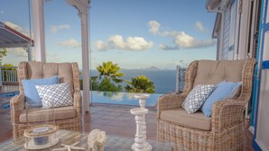A wicker lounge chair with a cushion and a decorative item on a glass table, overlooking a pool and a scenic view. - Luxury Villa in the French Carribean with a stunning view (Vieux Fort)