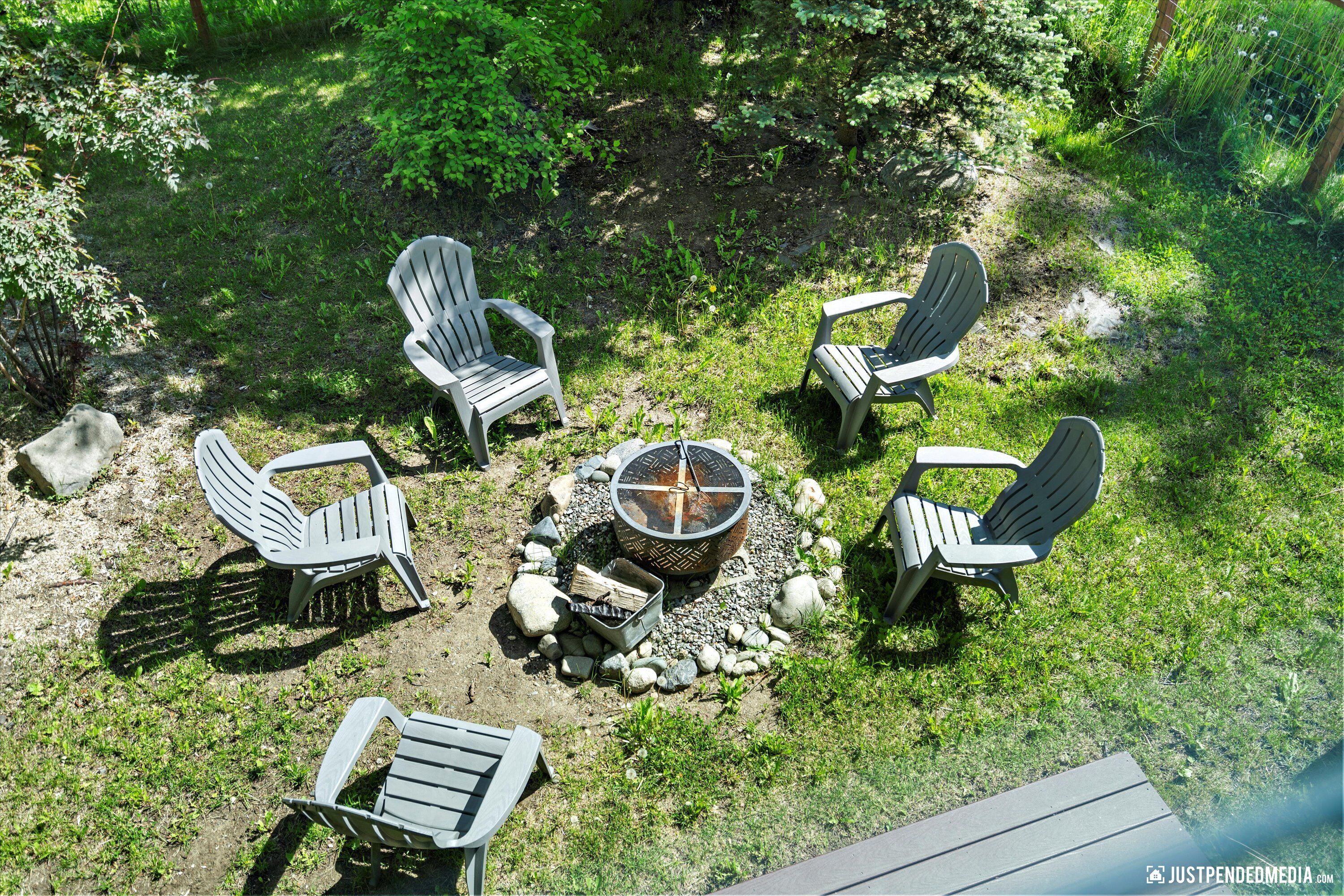 Disposición circular de asientos con sillas tipo Adirondack alrededor de un hogar central, ubicada en un área exterior con césped y árboles al fondo.
