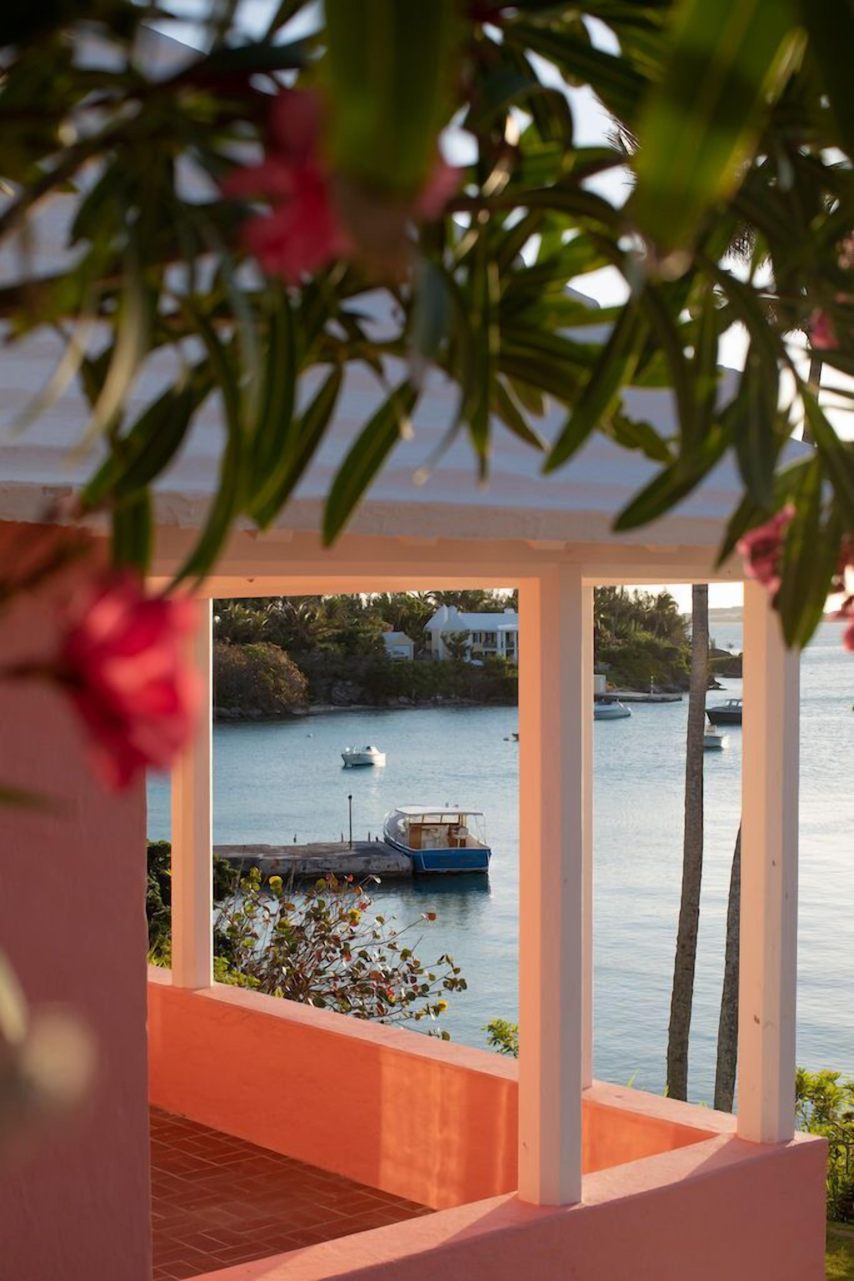 A view from a covered balcony overlooking a calm body of water with boats, palm trees, and a white building in the background.
