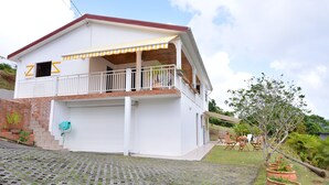 A two-story house with a balcony, a garage, and a garden area with potted plants. - Les Ixoras, quiet, flower-filled gîte in green setting 6 km from town and beaches. (La Trinité)