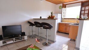 A compact living space with a TV, bar stools, a small kitchen, and a dining area with a table and chairs. - Les Ixoras, quiet, flower-filled gîte in green setting 6 km from town and beaches. (La Trinité)
