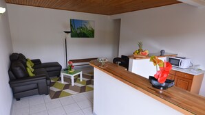 A modern living room with a sofa, a small table, and a kitchen area with a microwave and fruit bowl. - Les Ixoras, quiet, flower-filled gîte in green setting 6 km from town and beaches. (La Trinité)