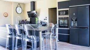 A modern kitchen with a black refrigerator, a microwave, and an oven. There are four clear plastic chairs around a central island.