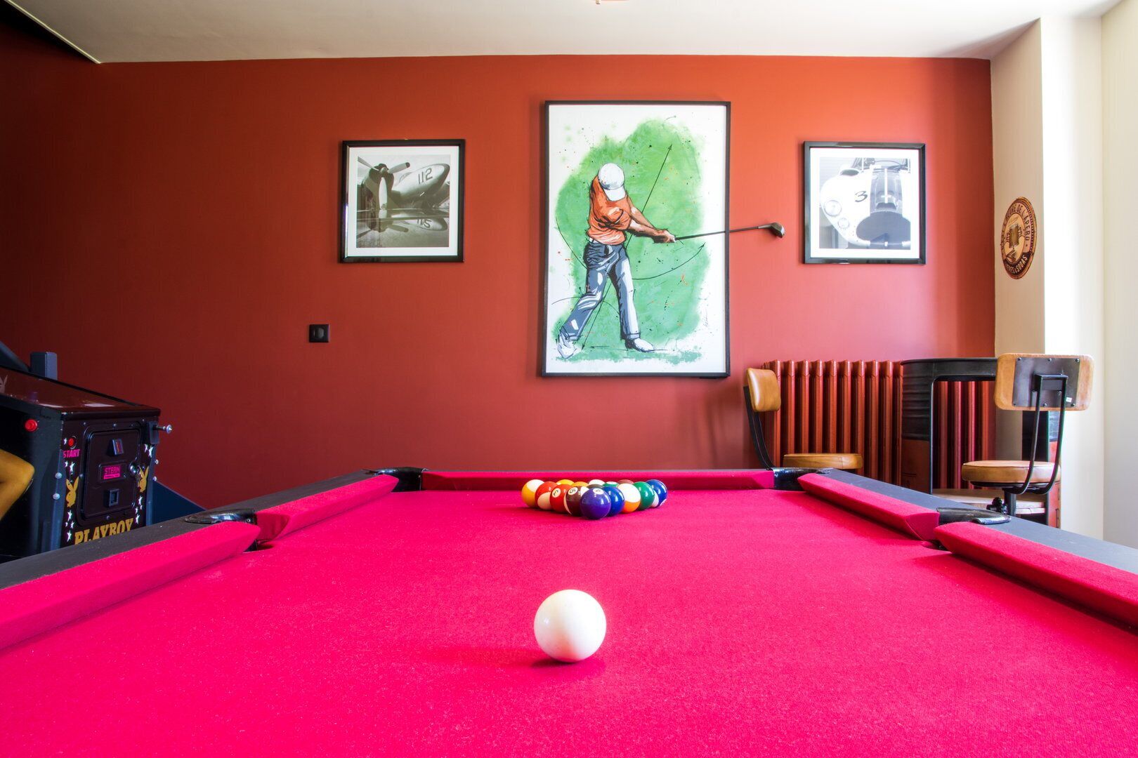 A pool table with a white cue ball, surrounded by colorful balls, in a room with red walls and two framed artworks depicting a vintage airplane and a golfer.