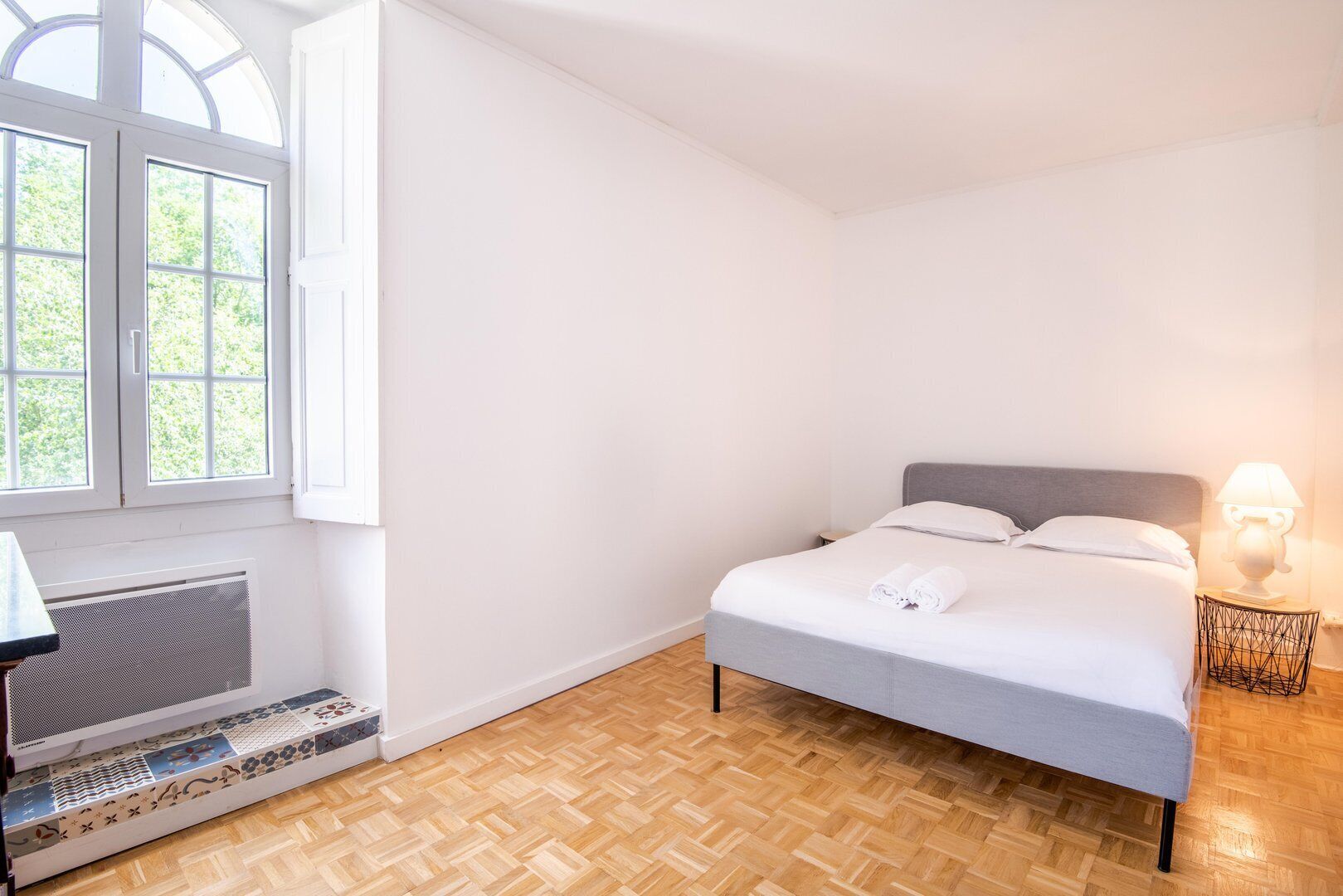 Un dormitorio con cama, mesita de noche, lámpara, radiador y ventana con vistas a zonas verdes.