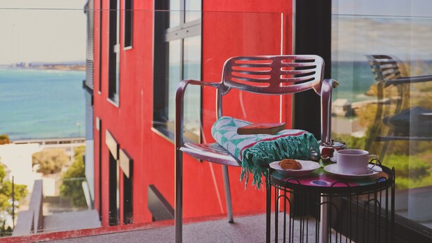 A balcony with a red building, a chair, a small table with a cup and saucer, and a view of the sea. - Redrock Ocean View Villa (Cape Town)