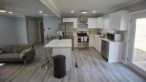 Une cuisine moderne avec des meubles blancs, des appareils en acier inoxydable, un îlot central et un coin repas agrémenté d’un canapé gris.