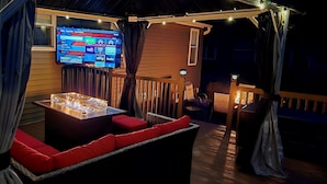 A cozy outdoor seating area with a fire feature, a flat-screen TV mounted on the wall, and a wooden deck. - The Silliman House - New renovation, Farmhouse Chic, close to baseball parks (maryland)