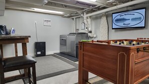Een kamer met een tafelvoetbaltafel, een wasmachine en een televisie waarop 'The Silliman House' staat.