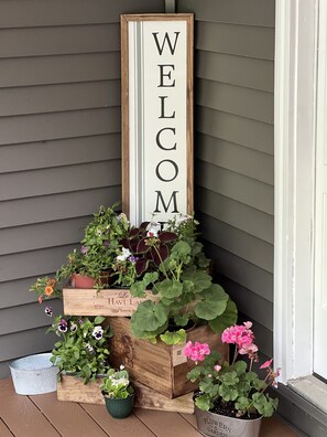 Een houten welkomstbord met de tekst 'WELCOME' op een veranda met verschillende potplanten.