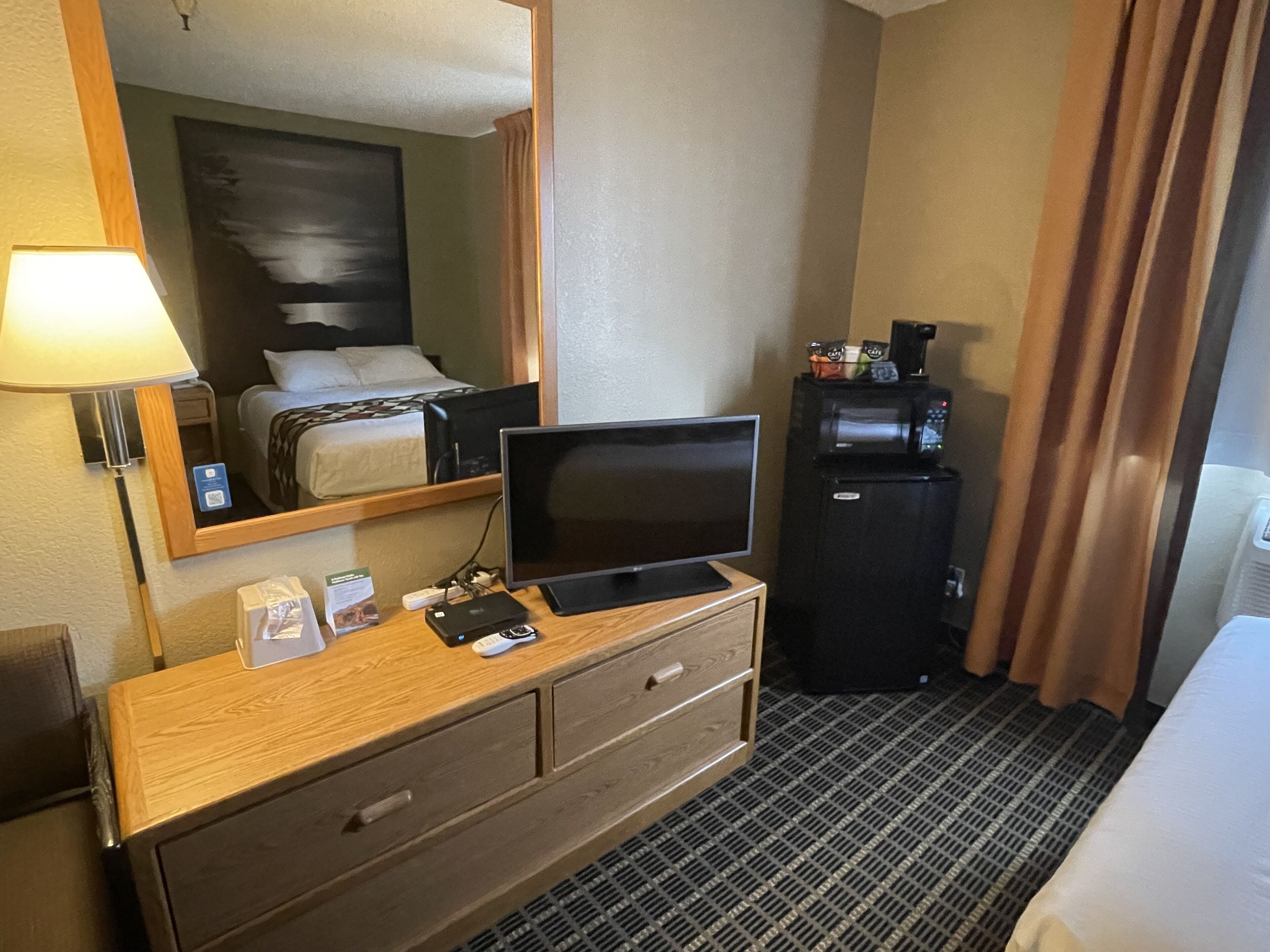 A hotel room with a wooden dresser, a flat-screen TV, a mini-fridge, a coffee machine, and a bed with a headboard.