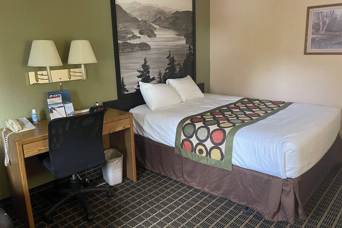 Une chambre d’hôtel avec un lit, un bureau, deux lampes, une chaise, une poubelle et un grand tableau représentant un paysage accroché au mur.