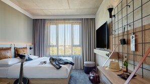 Una habitación de hotel con una cama, una guitarra, un televisor, un estante montado en la pared con una botella y un sombrero, y una ventana con cortinas.