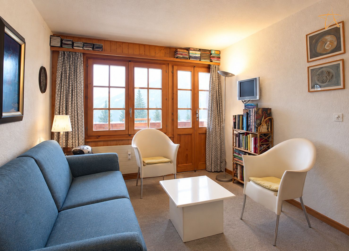A living room with a blue sofa, two white chairs, a white coffee table, a bookshelf, a television, and a window with curtains.