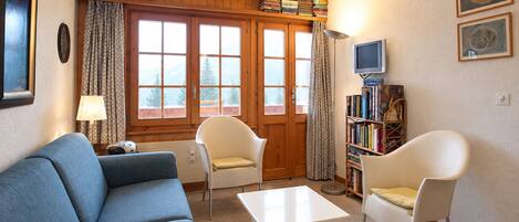 A living room with a blue sofa, two white chairs, a white coffee table, a bookshelf, a television, and a window with curtains.