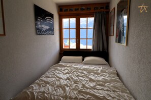 A narrow room with a bed, a window with curtains, and a wall with a framed picture and a sign. - Cosy apartment with south-facing balconyOutside : apartment on the 2nd floor with south-facing balco (St-Luc)
