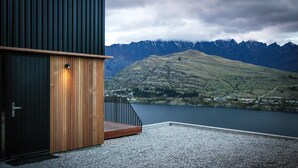 A modern building with a wooden door and a staircase leading to a gravel area overlooking a lake and mountains.