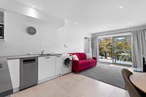 A modern kitchen with a dishwasher, washing machine, and a red sofa near a sliding glass door leading to a balcony with a view of trees and hills.