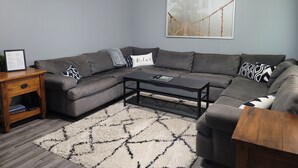 A modern living room with a grey sectional sofa, a coffee table, a wooden side table, and a framed picture on the wall. - Comfortable 4 Bedroom / 1.5 Bathroom Home (Ypsilanti)
