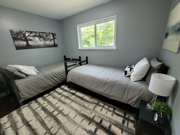 A bedroom with two single beds, a window with a view of greenery, a painting on the wall, and a bedside table with a lamp and a clock. - Comfortable 4 Bedroom / 1.5 Bathroom Home (Ypsilanti)