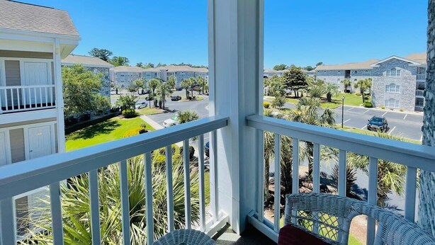 A balcony with white railings, wicker furniture, and a view of a residential area with multiple buildings and palm trees. - Great 1 BDR Condo in the center of Myrtle Beach (Myrtle Beach)