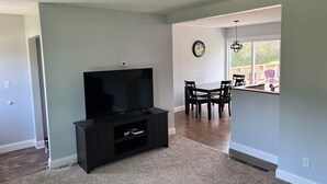 A living room with a flat-screen TV on a black stand, a dining area with a table and chairs, and a large window with a view of a deck. - Ranch home with 3 bedrooms upstairs and 1 down. Refurnished June of 2022 (St. Ignace)