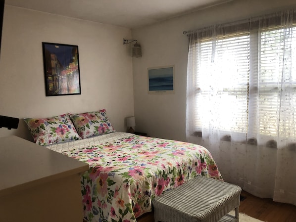 Un dormitorio con cama, mesita de noche, lámpara, banca de mimbre y ventana con cortinas traslúcidas.