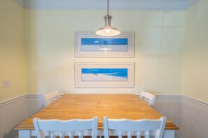 A dining room with a wooden table and white chairs, framed artwork on the wall, and a hanging light fixture.