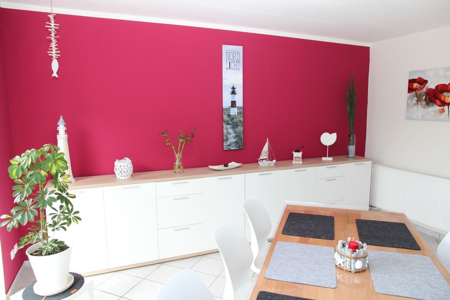 Ein Essbereich mit roter Wand, weißen Schränken und einem Holztisch mit grauen Untersetzern.