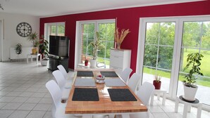 Un comedor con mesa de madera y sillas blancas, ventanas amplias con vista a zonas verdes, un televisor y plantas en macetas.