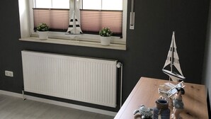 Una habitación con un ventanal grande, un radiador debajo y una mesa de madera con un modelo de velero y una botella.