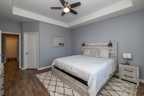 A bedroom with a bed, a nightstand, a ceiling fan, a door, and a hallway.